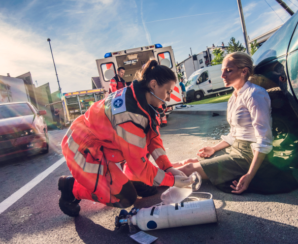 A paramedic tends to a woman's injured leg beside a car accident site on a sunny day, with an ambulance and another paramedic visible in the background.