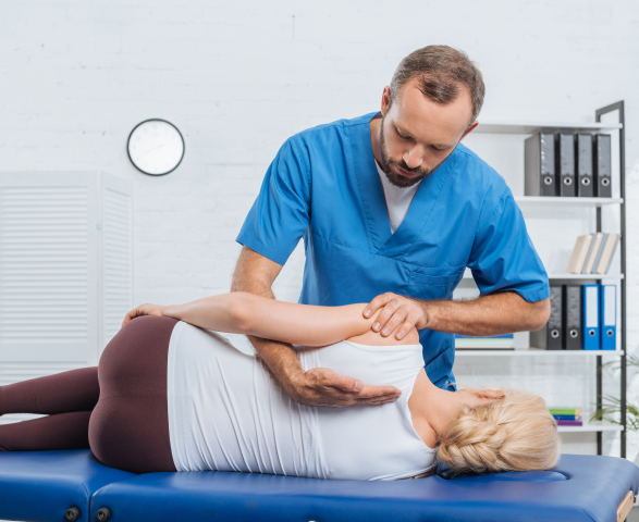 A chiropractor in blue scrubs carefully adjusts a female patient's back as she lies on a treatment table in a clinic with a white brick wall and shelves in the background.