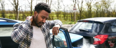 A man standing by his car, which has a visibly damaged rear end and smoke emitting from it from a recent car accident, appears stressed and is holding his neck in discomfort.