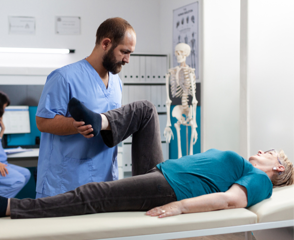A physical therapist assists an elderly female patient with leg exercises in a clinic to manage joint pain, with a skeletal diagram on the wall in the background.