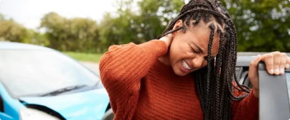 A young woman with braided hair, wearing a rust-colored sweater, winces and holds her neck, standing beside a blue car after a car accident, possibly experiencing neck pain or distress.