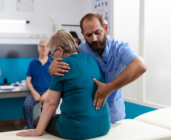 A physical therapist assists an elderly woman with an exercise, gently supporting her back for spine health in a clinical setting, while another patient observes in the background.
