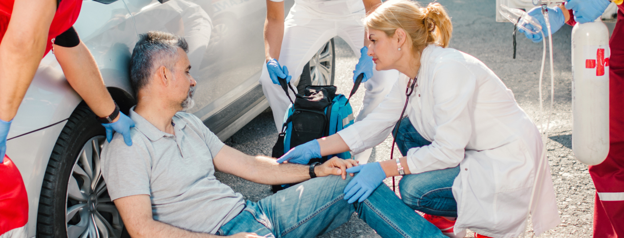 A paramedic provides first aid to an injured man sitting on the ground by a vehicle, with another rescuer looking on. They are outdoors, equipped with medical supplies.