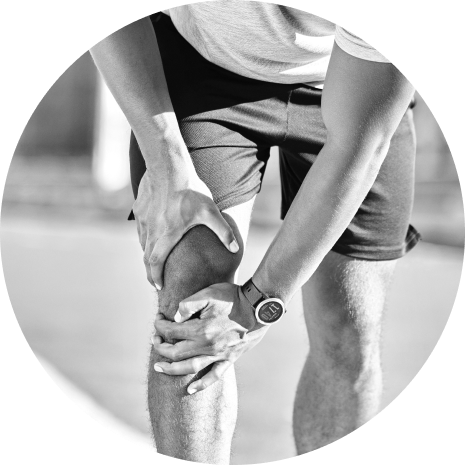 A close-up image in black and white of a person holding their knee, possibly indicating joint pain or injury, while wearing shorts and a wristwatch.