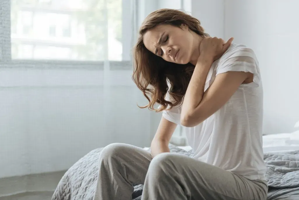 A woman sitting on the edge of a bed, holding her neck in pain with a grimace, indicating discomfort or a potential injury, in a bright room. This could prompt one to wonder why neck