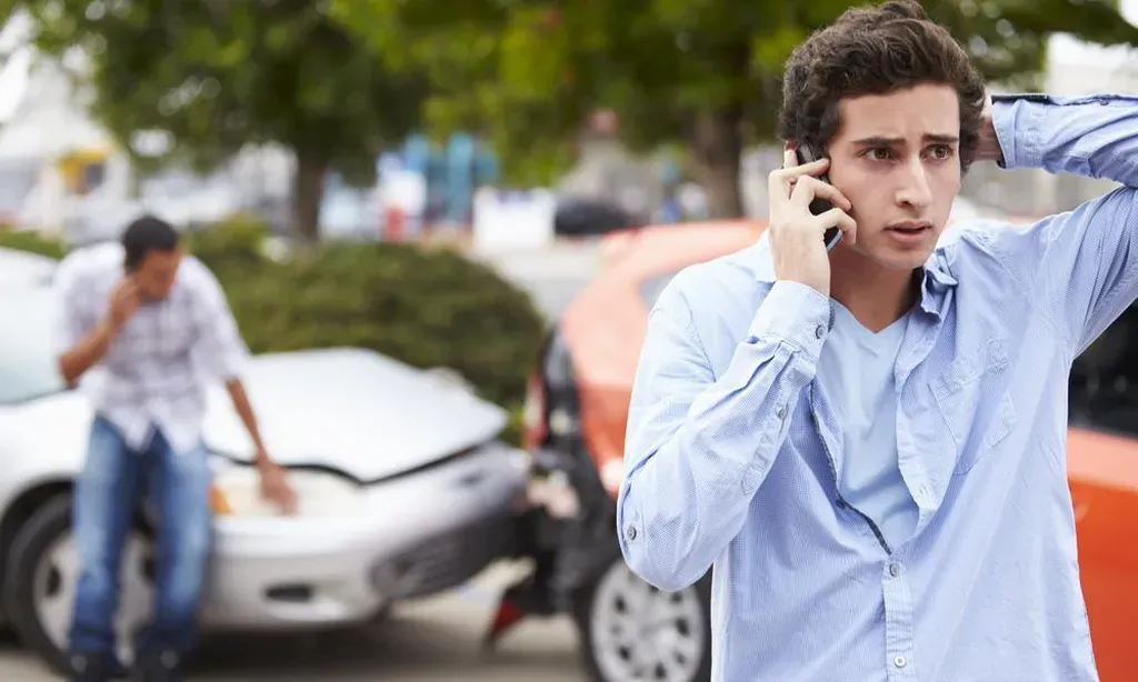 A worried young man talking on a cell phone with his hand on his head, standing near an auto accident scene where another man is checking a damaged vehicle hood.