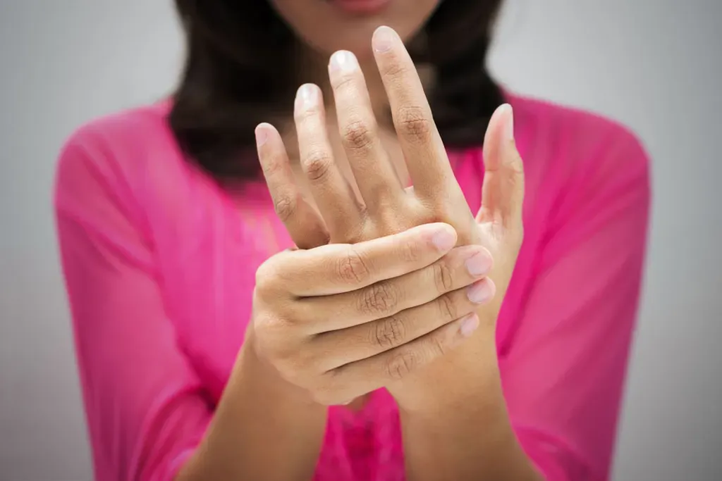 A woman in a pink blouse holds her wrist, possibly experiencing early signs of arthritis. The focus is on her hands, with the background softly blurred.