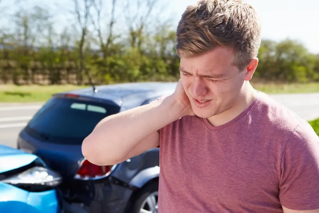 A distressed young man holds his neck in pain in front of two collided cars on a sunny day, indicating he might be experiencing whiplash or other car accident injuries.