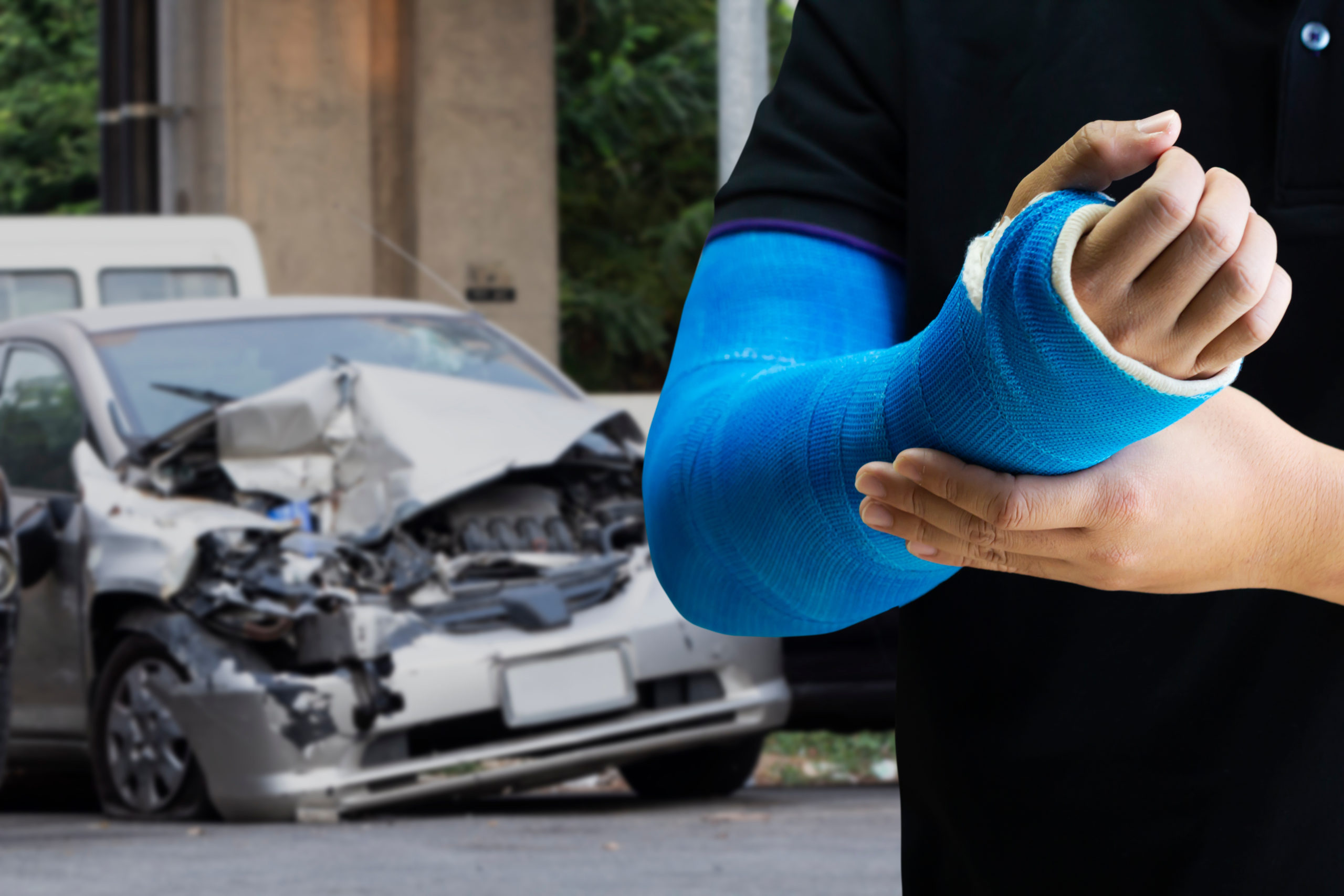A person in a black shirt wearing a blue cast on their forearm, with a heavily damaged car in the background, implying an auto accident injury.