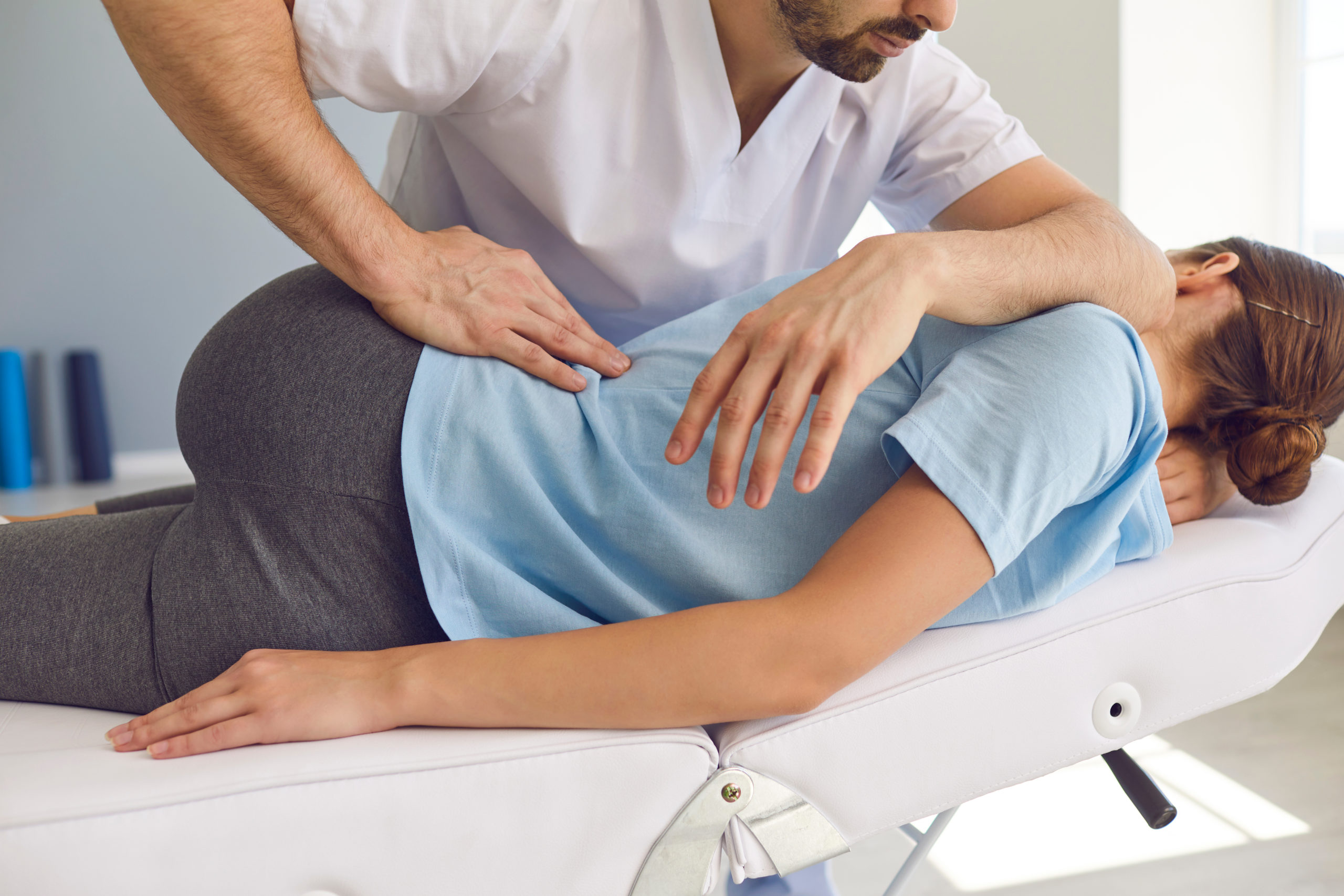 A person receiving a chiropractic adjustment while lying on their side on a white cushioned table. The chiropractor, dressed in a white shirt, is applying pressure to the person's lower back with one hand and supporting their arm with the other, promoting spinal health through the science of chiropractic.