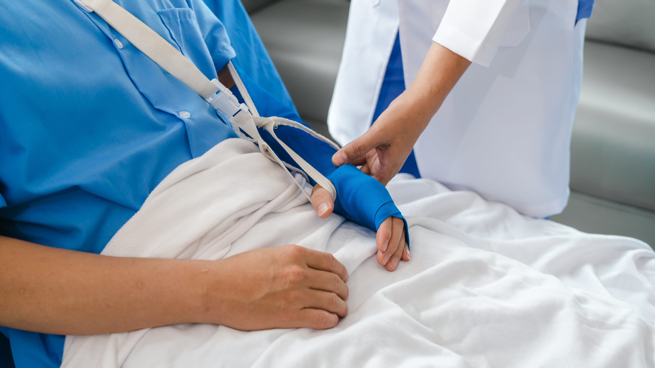 A person lies in a hospital bed with a blue sling supporting their right arm, highlighting the importance of proper auto injury treatment. A healthcare professional adjusts the sling, ensuring correct support. The patient wears a blue hospital gown and is covered with a white blanket, illustrating fact vs. myth in recovery care.