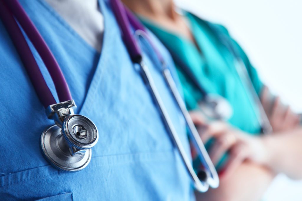 Close-up of two medical professionals in scrubs, one wearing a stethoscope around the neck, focused on enhancing recovery. The background is a soft blur, emphasizing their upper torsos and the medical equipment.