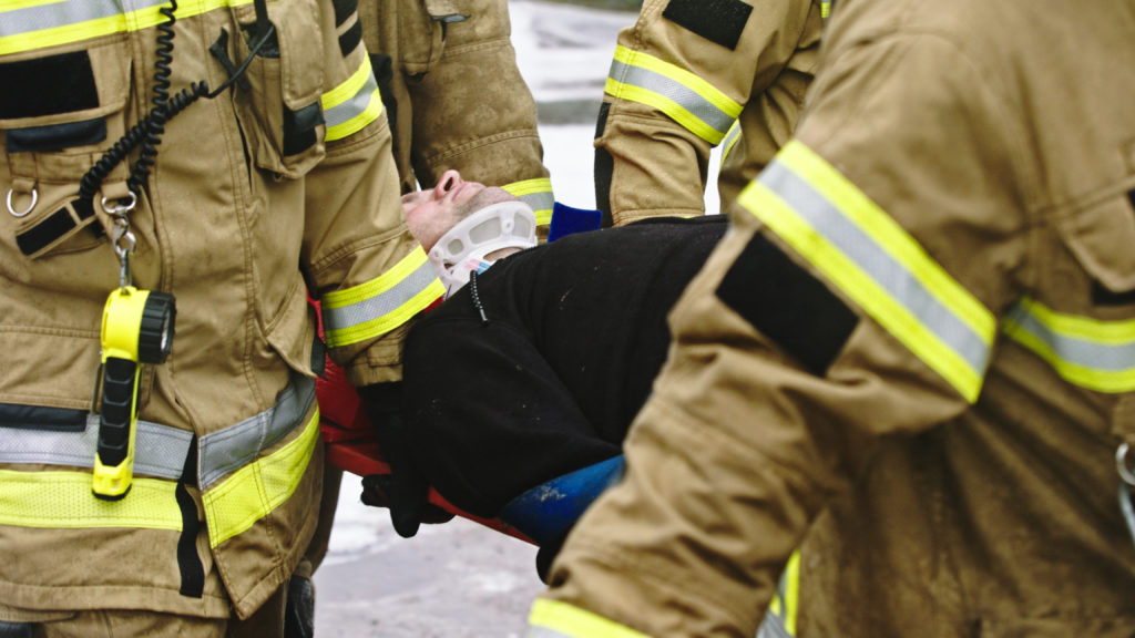 Three firefighters in full gear are carrying an injured person on a stretcher, likely due to car accident injuries. The person is wearing a cervical collar for neck support and is partially visible, lying on their back. The scene clearly depicts the urgent impact such incidents can have on daily life.