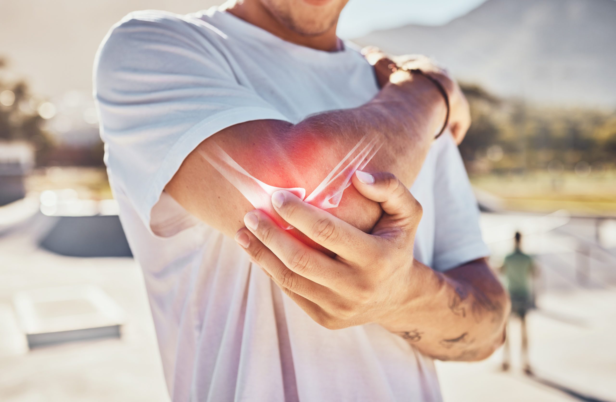 A man wearing a white T-shirt holds his right elbow with his left hand outdoors, showing signs of serious joint pain. A red glow around the elbow indicates injury. In the blurred background, there is a mountain and a person standing in a park.