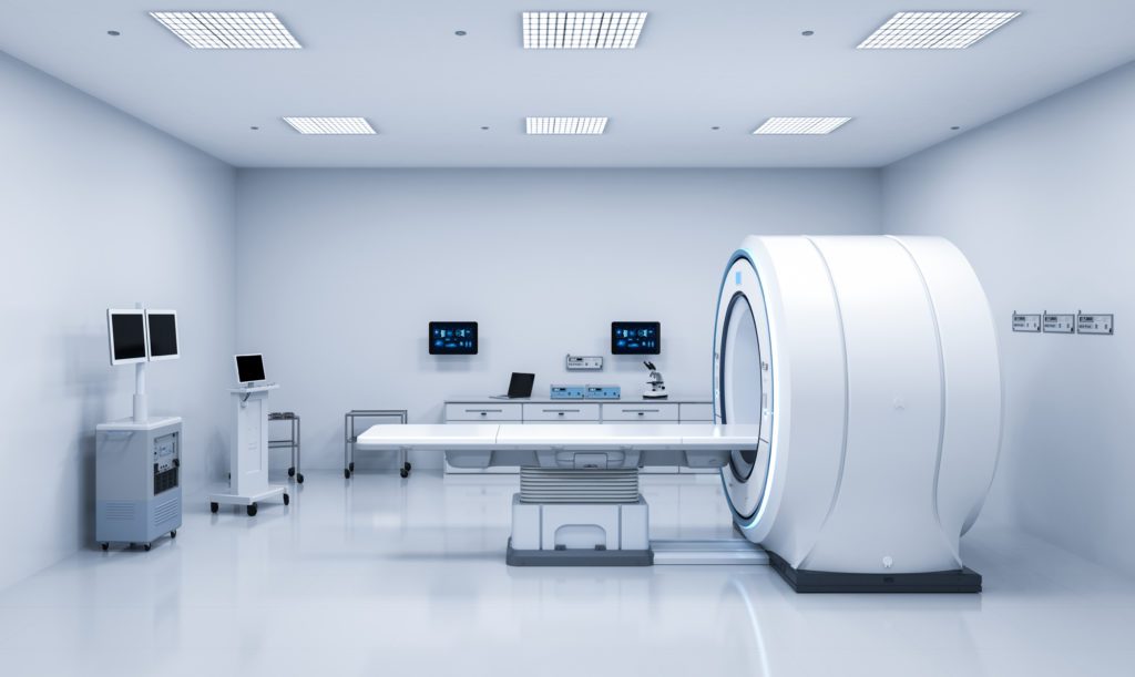 A modern, white-walled medical imaging room contains a large MRI machine with a patient table, multiple computer monitors mounted on walls, a trolley, and medical equipment on mobile stands. For a smooth experience during your imaging appointment, the room is brightly lit with ceiling lights.