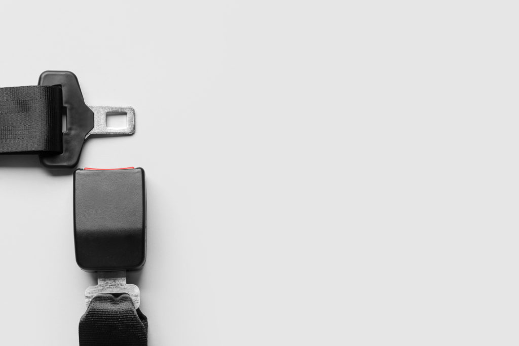 A close-up image of a black car seatbelt against a light gray background. The seatbelt buckle and latch plate are positioned next to each other but not connected. The latch plate is on the left, and the buckle is on the right, highlighting their role in preventing car accidents and seat belt injuries.