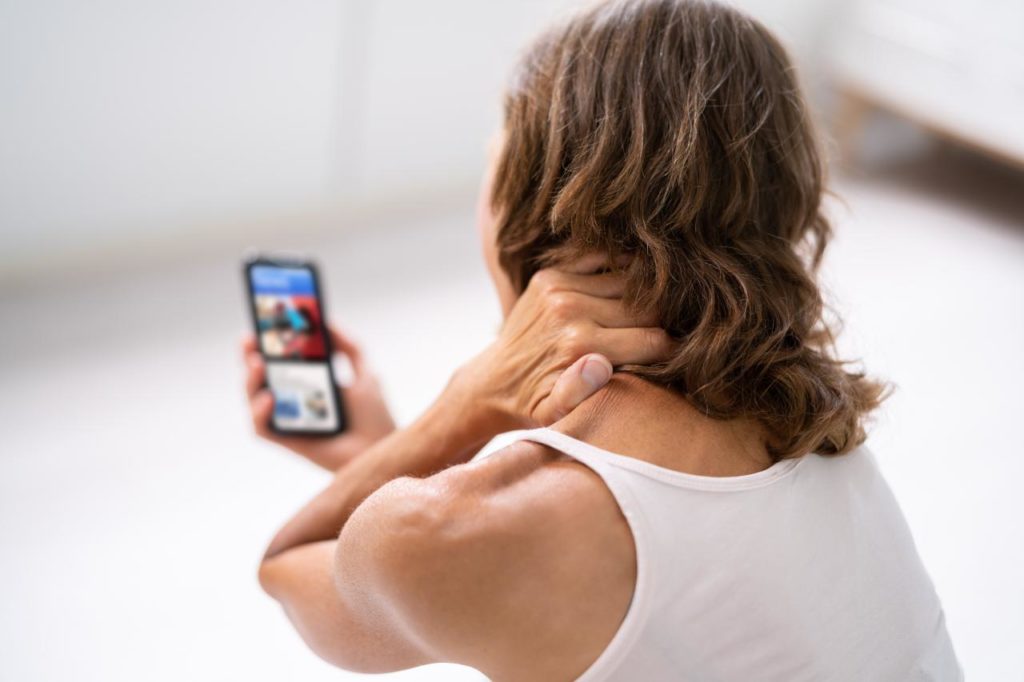 A person with shoulder-length brown hair, wearing a white sleeveless top, is rubbing their neck with one hand while looking at a smartphone held in the other. The image shows the person's back and part of their profile, suggesting neck pain or tension.