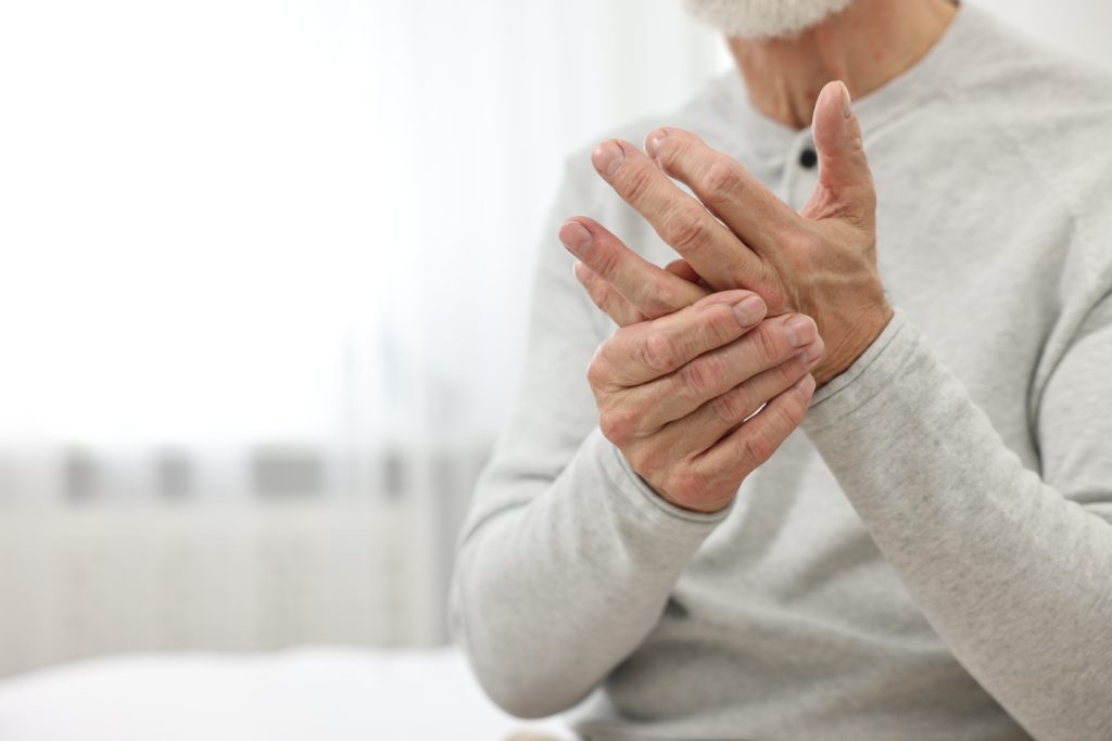 A person wearing a gray long-sleeve shirt is sitting and massaging their hand, possibly experiencing joint pain or discomfort. The background is blurry with a soft, bright indoor setting. The person's face is not visible in the image, debunking the myth that weather changes cause joint pain.