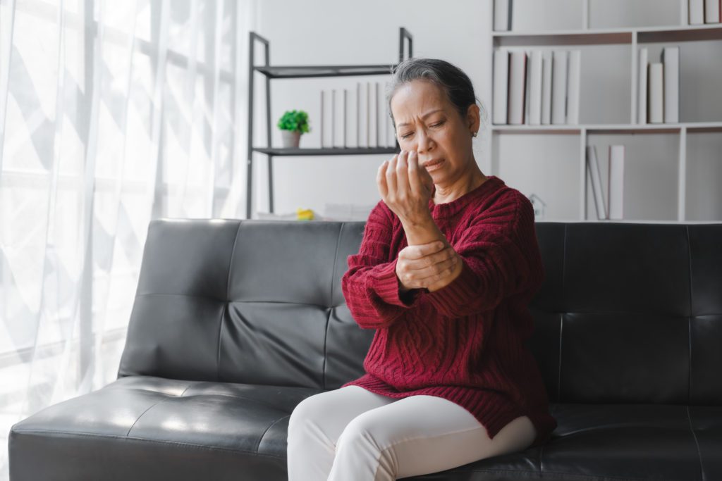 A woman is sitting on a black couch in a well-lit room, wearing a red sweater and white pants. She is holding her right wrist with a pained expression on her face, likely due to migratory arthritis. In the background, a shelf with books and a green plant is visible.