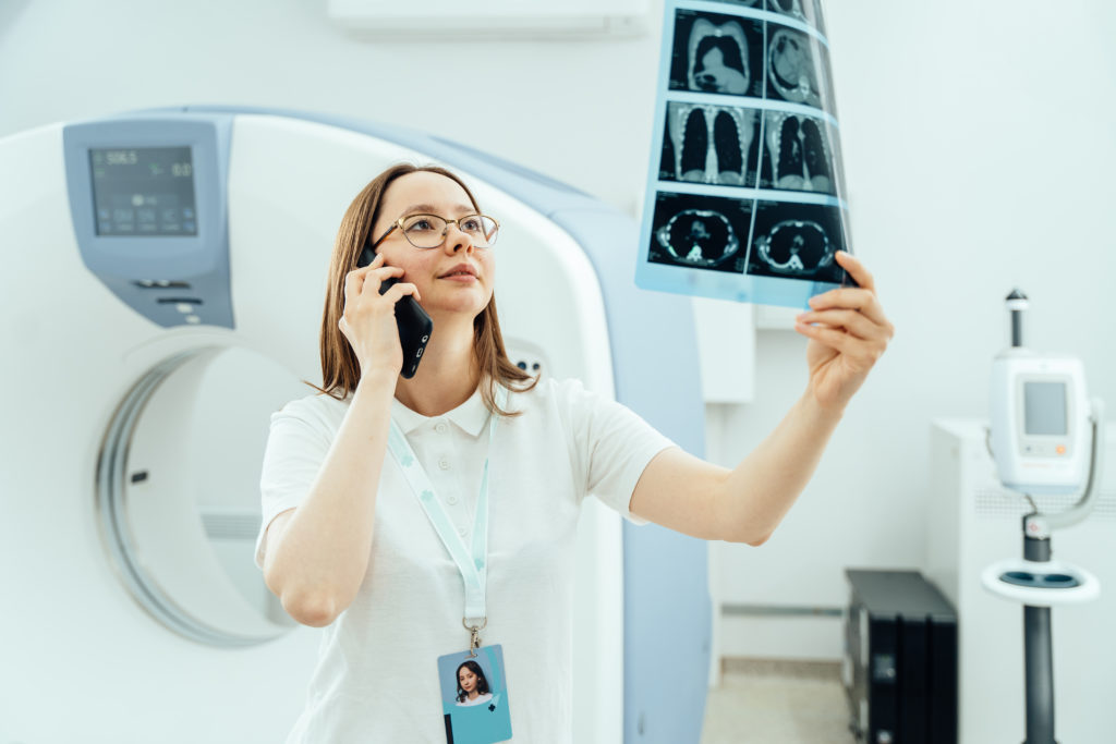 A medical professional wearing glasses and a white coat, holding an anatomically labelled ID card around their neck, speaks on the phone while examining patient X-ray images in a medical imaging room. A CT scanner used for whole body scans and other medical equipment are visible in the background.