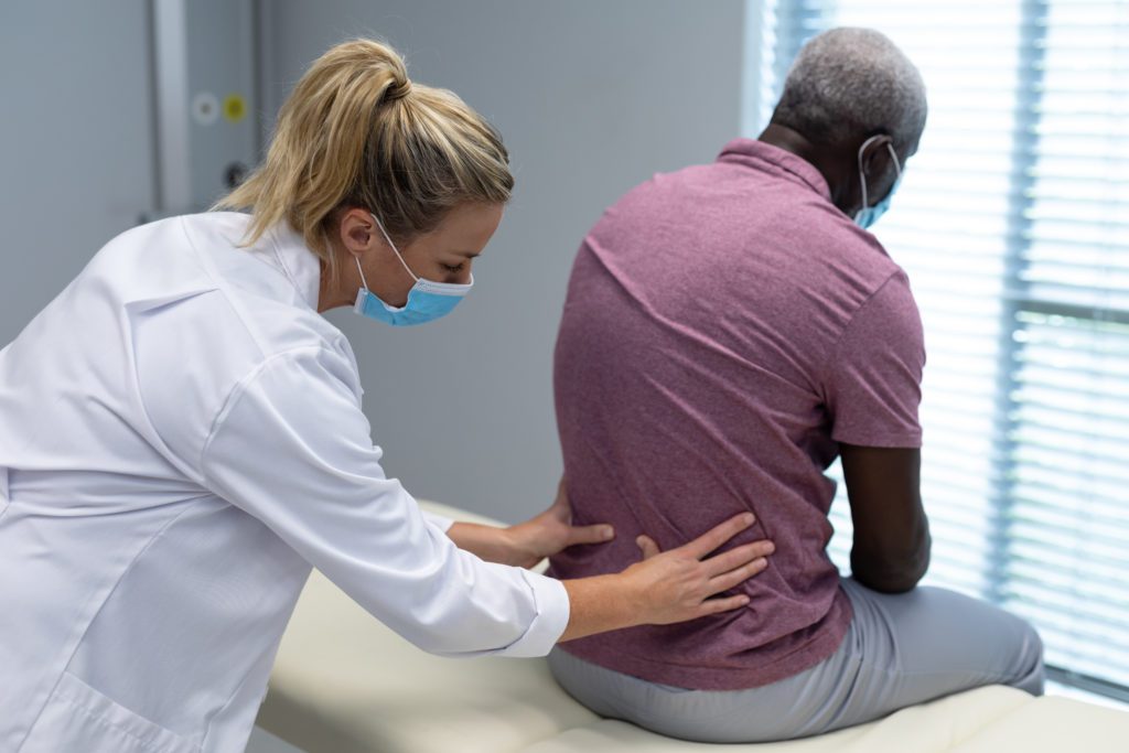 A healthcare professional wearing a face mask examines an elderly man who also wears a face mask. She uses her hands to check his lower back for causes of back pain as he sits on an examination table. The room is well-lit with natural light coming through window blinds.