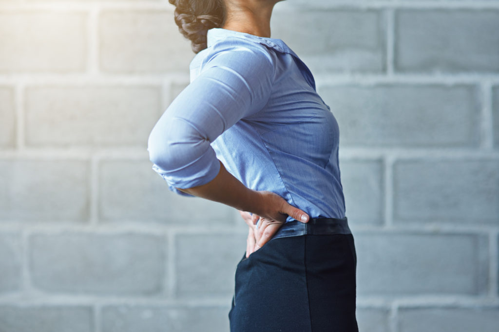 A person in a blue shirt and black pants stands against a gray brick wall, arching their back and placing both hands on their lower back, as if experiencing everyday back pain.