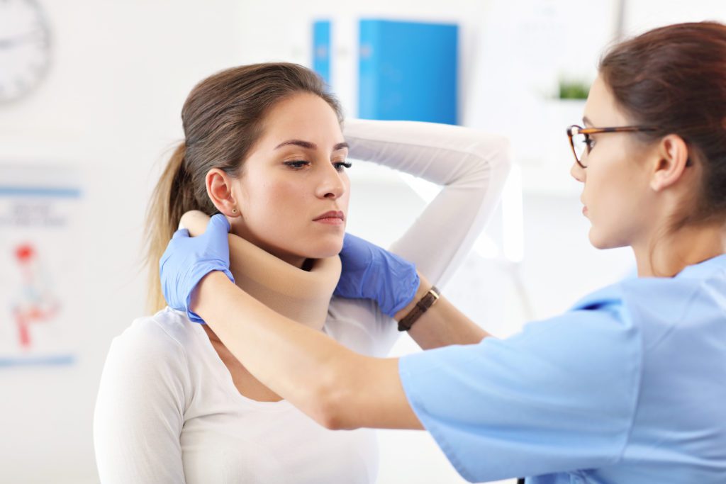 A medical professional in blue scrubs and gloves is adjusting a neck brace on a patient with brown hair who appears to have neck problems. The patient, possibly experiencing symptoms from a car accident, is in a white top and sitting up straight, while the medical professional focuses on fitting the brace properly.