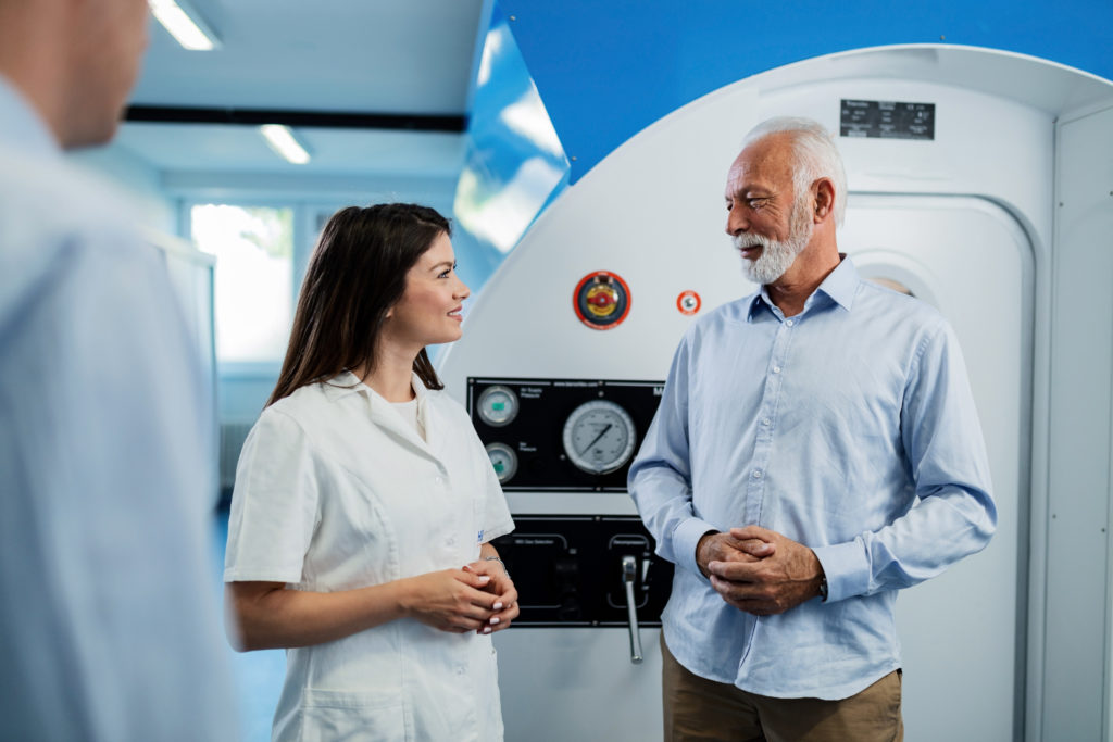 Happy female doctor and senior man communicating about therapeutic effects of hyperbaric chamber treatment.