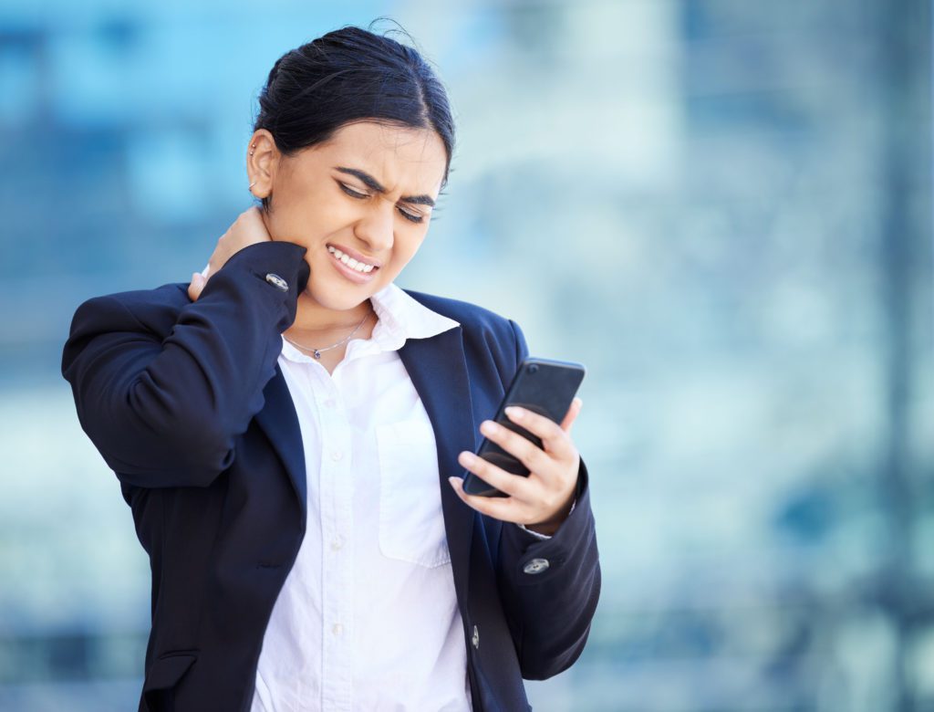 A person in a business suit stands outdoors, squinting in discomfort while holding a smartphone and rubbing their neck. The background is blurred, suggesting an urban environment. The person appears to be experiencing cell phone neck while using their phone.