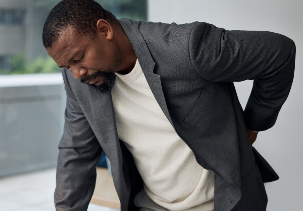 A man in a grey suit and white shirt grimaces in pain, holding his lower back with one hand while leaning forward. He appears to be experiencing everyday back pain, standing near a window with a blurry background.