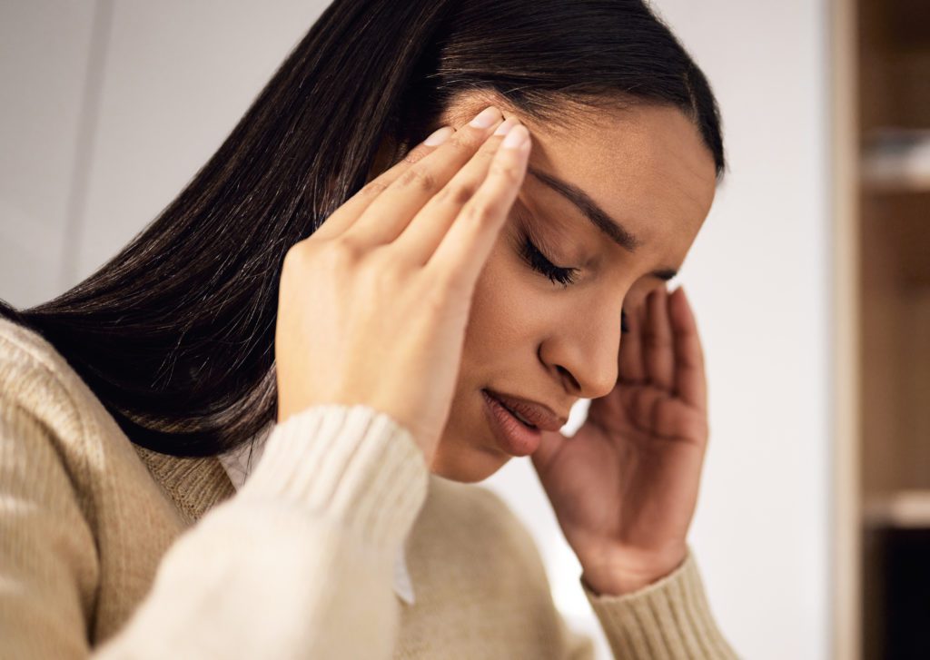 A woman with long dark hair and dressed in a beige sweater is holding her temples with both hands, eyes closed, and appears to be experiencing a headache or stress. Consider chiropractic adjustments for potential health benefits to improve your health. The background is out of focus.