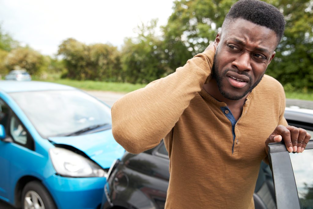 A man stands beside a car, holding his neck in apparent pain, suggesting possible neck problems. Two cars are shown in the background—one blue car with a damaged front and one black car—indicating a recent car accident. Green trees and a cloudy sky are visible in the background.