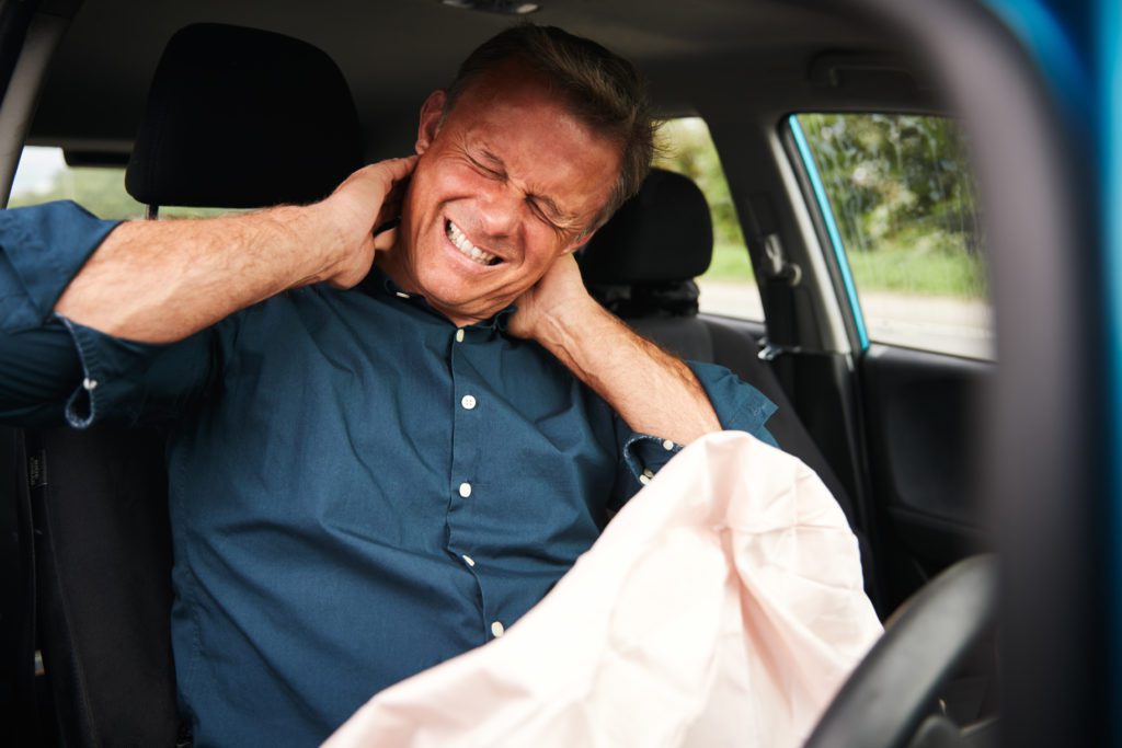 A man in a blue shirt sits in a car, grimacing in pain and holding his neck with both hands. The deployed airbag suggests he was just in a car accident. The blurred greenery outside adds context, hinting he's seeking solutions for his sudden neck problems.