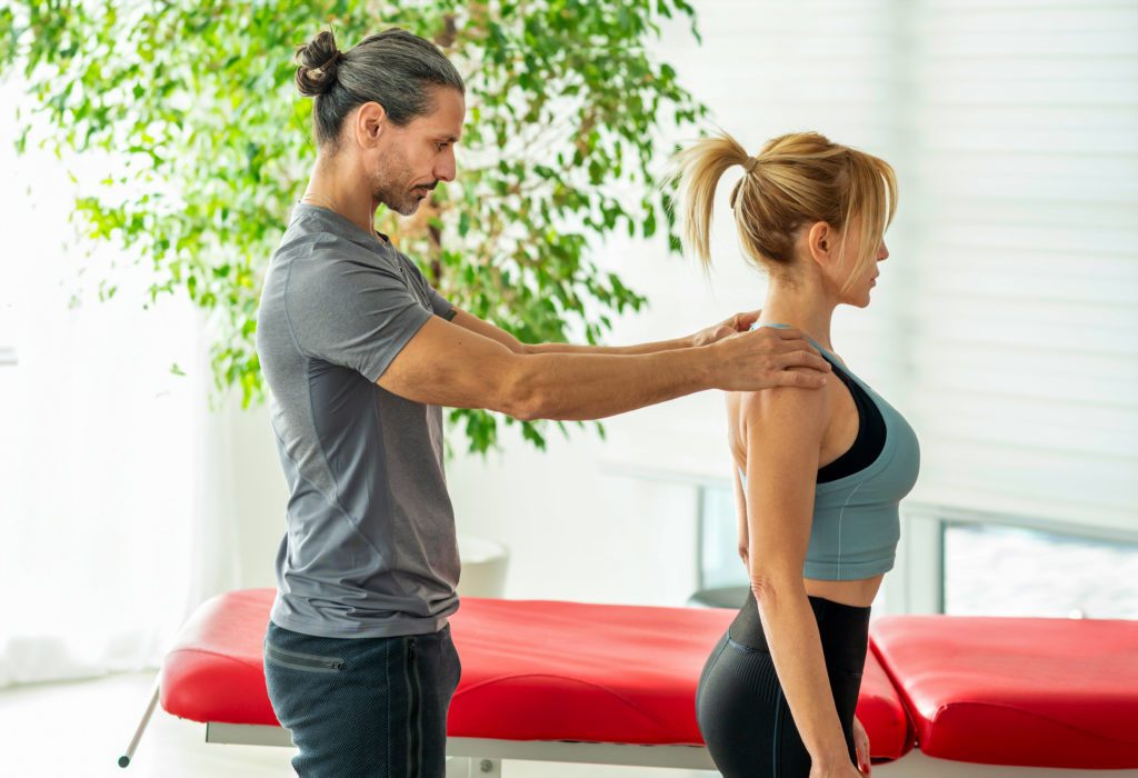 A bearded man with a bun in casual sportswear assists a blonde woman in a blue sports bra and black leggings by adjusting her shoulders. They are in a bright room with a large plant and a red padded table, highlighting the health benefits of chiropractic adjustments.