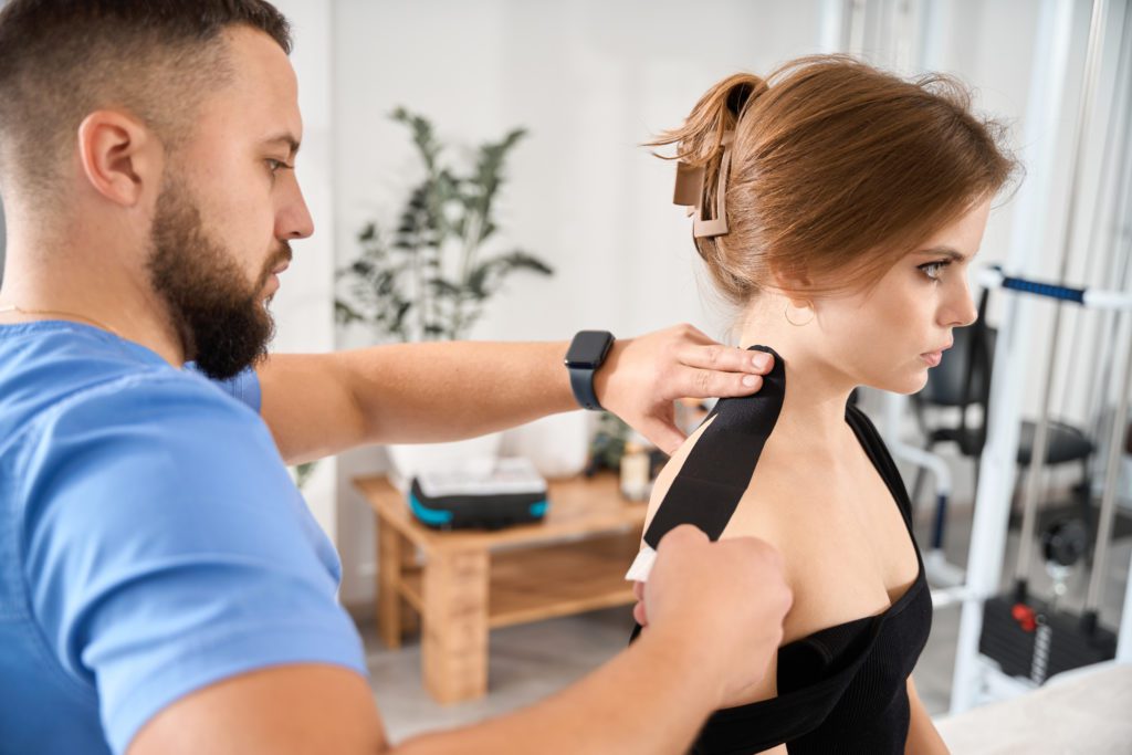 Man puts black tape on a patients shoulder, a woman in comfortable black clothes