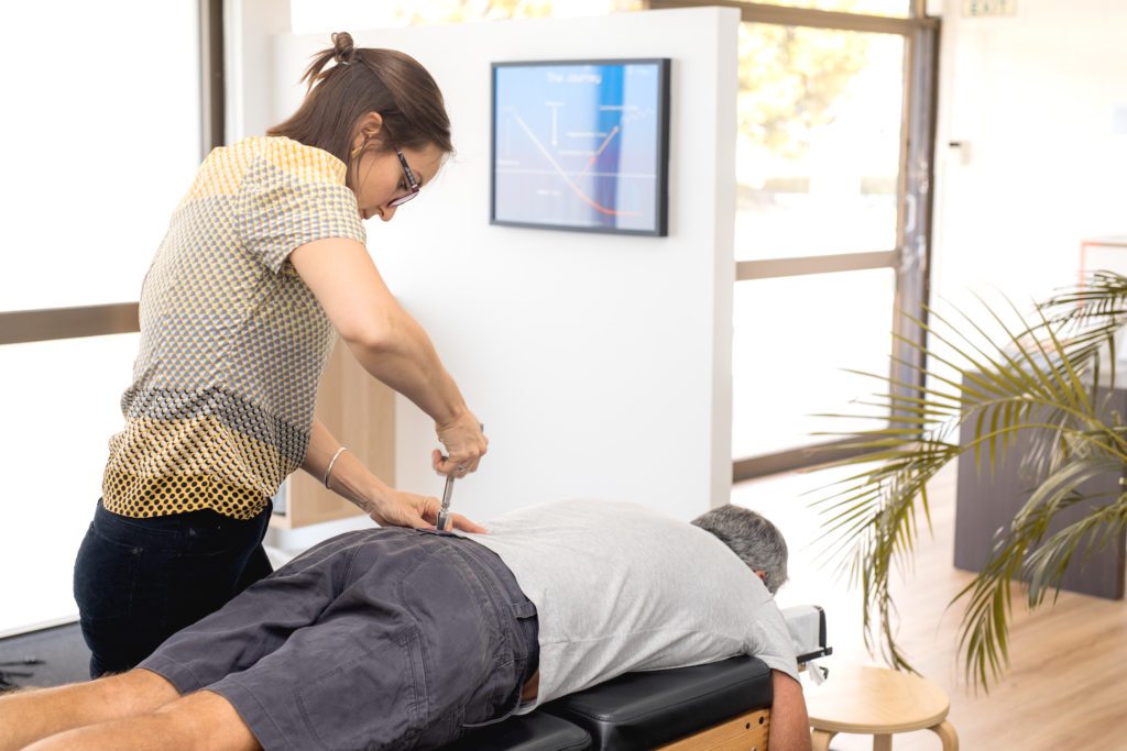 A chiropractor wearing glasses uses an activator tool on a patient's lower back. The patient, seeking relief from everyday back pain, lies face down on a chiropractic table in a brightly lit room with large windows, a plant, and a wall-mounted screen displaying a chart.