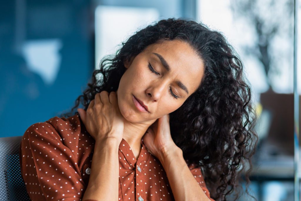 Close-up photo. Tired young hispanic woman. She sits in the office, holds and massages her neck with her hands, closed her eyes, feels pain, tension. Long working overtime day, deadline.