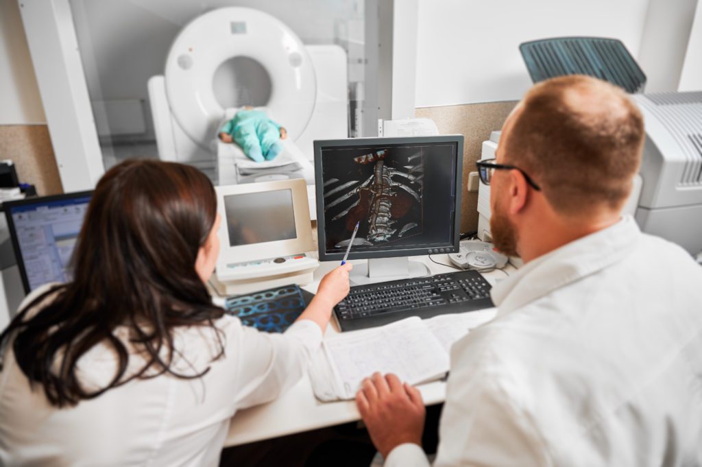 Two medical professionals analyze a CT scan on a computer monitor in a hospital room. A patient lies inside a CT scanner in the background. One professional points at the scan, discussing the advantages and potential disadvantages of whole body scans. They are surrounded by medical equipment and screens.